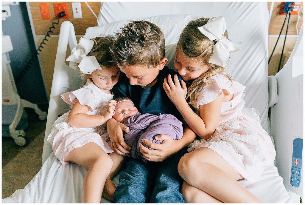 Siblings holding newborn baby in hospital bed